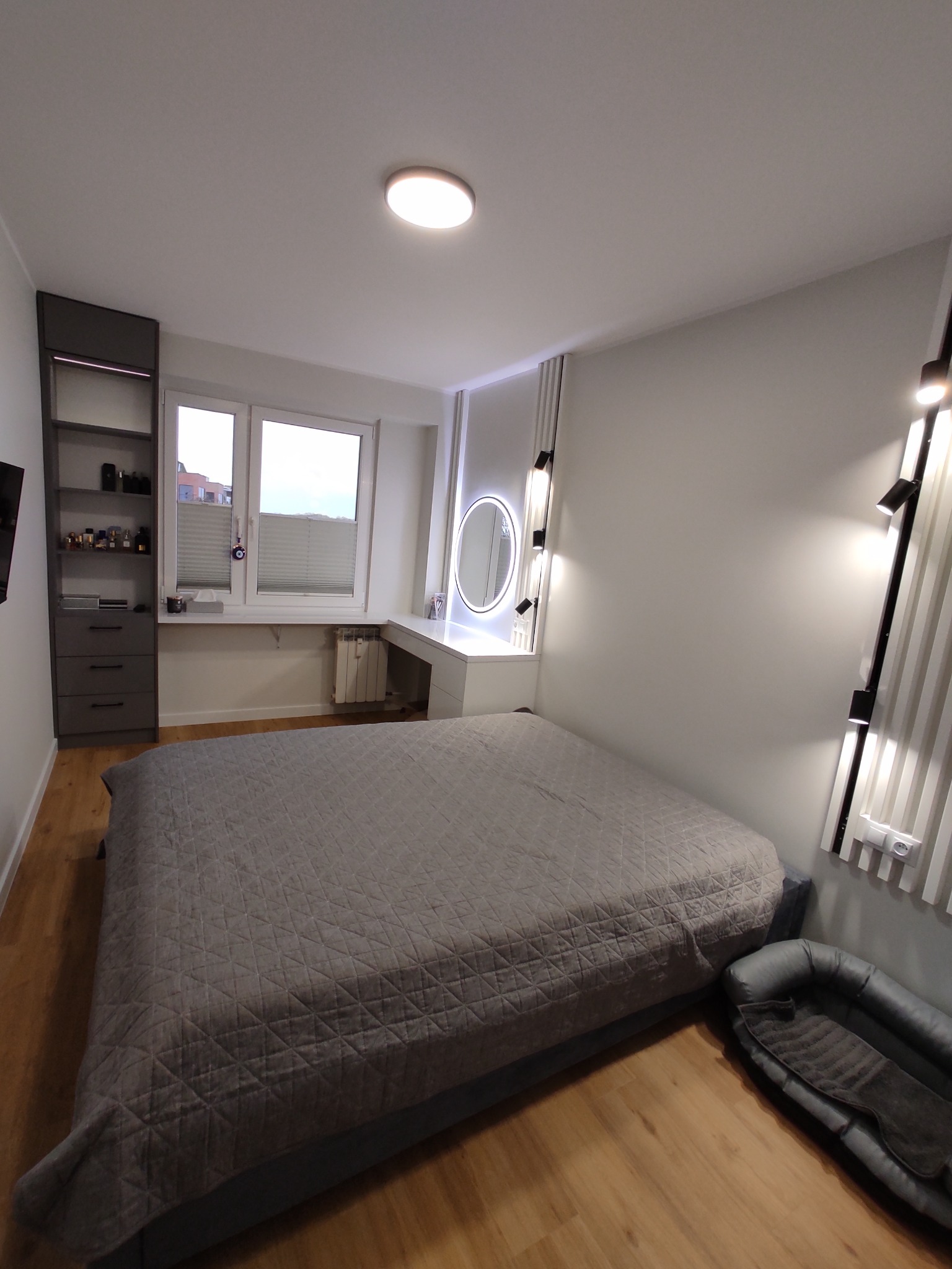 Nowoczesna sypialnia z szarą pościelą w geometryczne wzory, oświetleniem punktowym na ścianie i toaletką z okrągłym lustrem podświetlanym LED, uzupełniona o legowisko dla psa w rogu pomieszczenia.