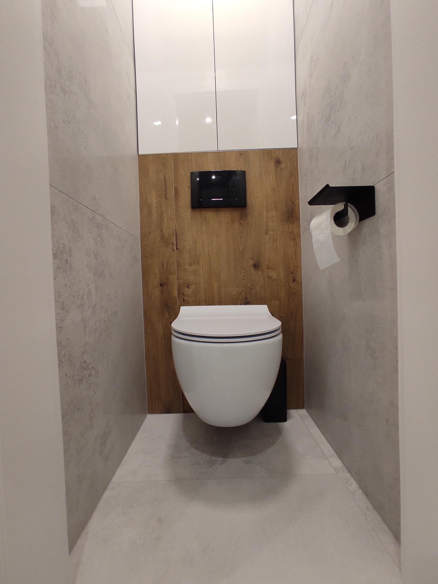 Nowoczesna toaleta podwieszana z drewnianym panelem i szarą płytką imitującą beton, minimalistyczny design, czarny uchwyt na papier toaletowy.