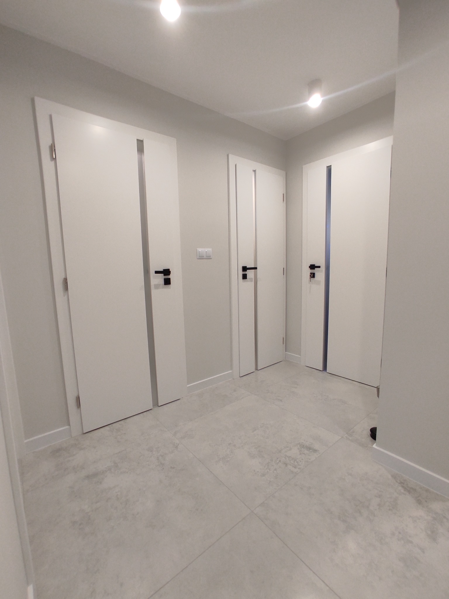 Jasne wnętrze korytarza z trzema białymi drzwiami z czarnymi klamkami, szara podłoga z płytek, oświetlenie sufitowe.