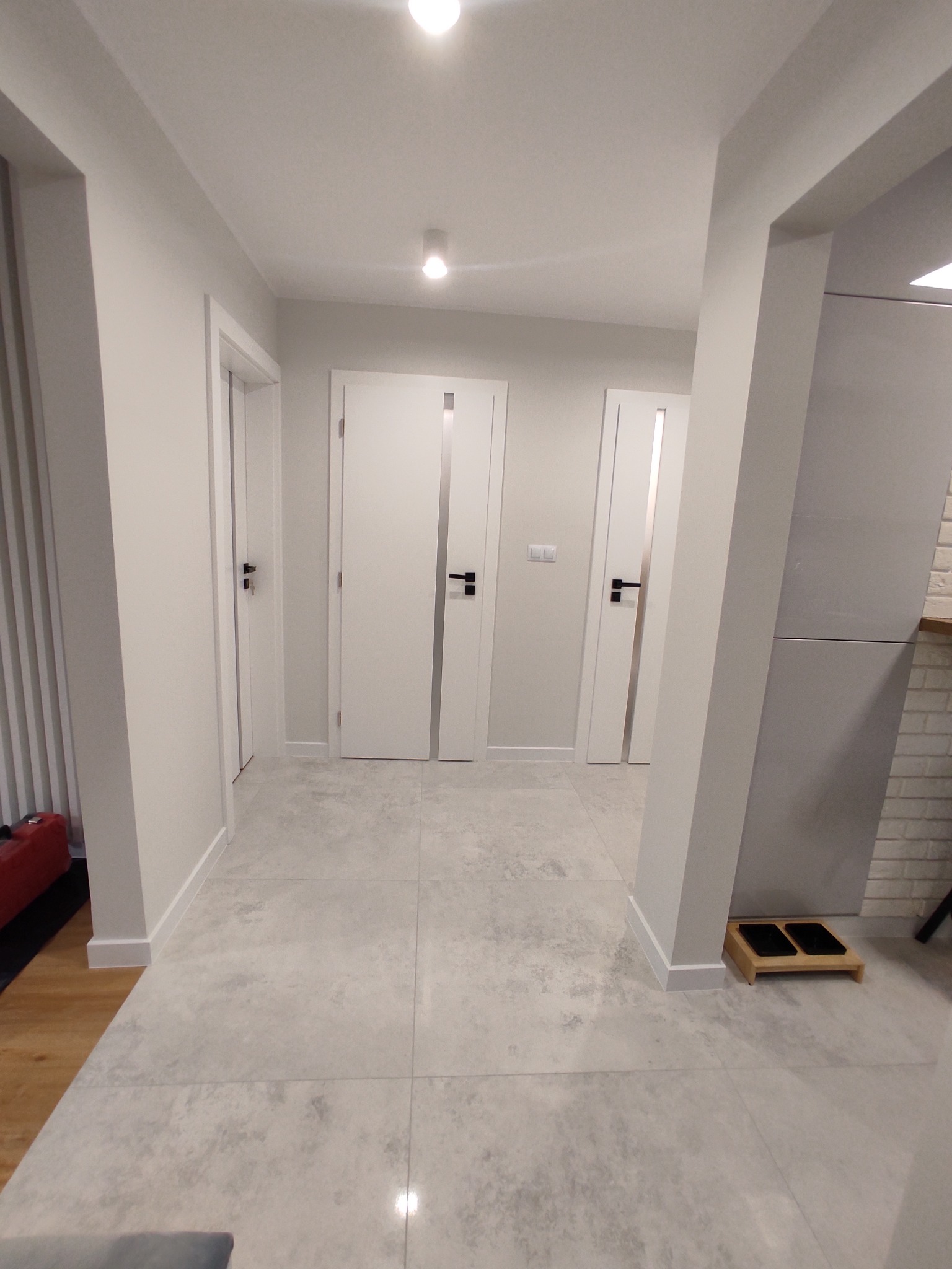 Nowoczesny korytarz w odcieniach bieli i szarości z trzema parami drzwi z czarnymi klamkami i szklanymi wstawkami, wykończony płytkami podłogowymi imitującymi kamień, oświetlony punktowo.