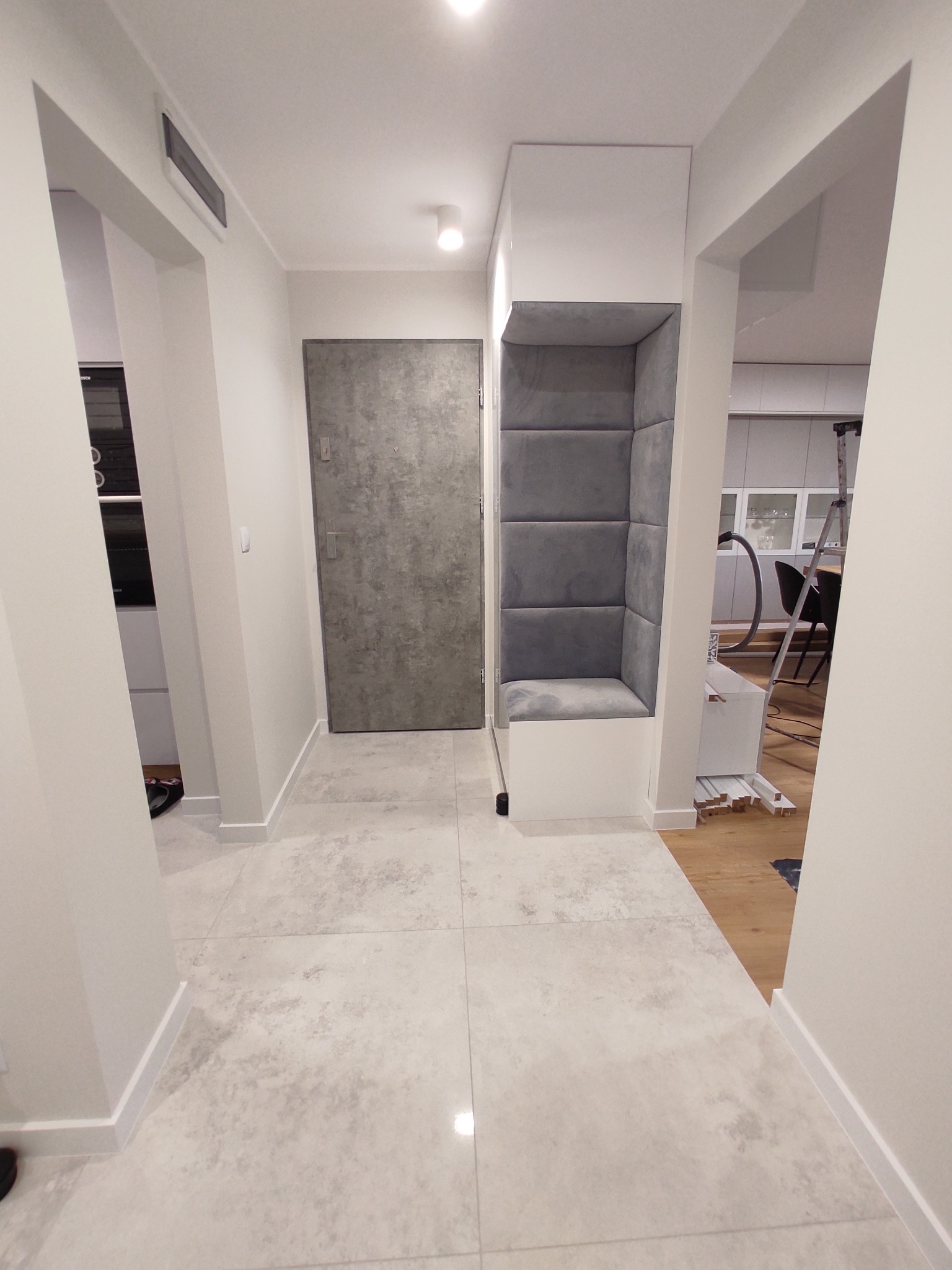 Wnętrze korytarza z zabudowaną wnęką z szarym tapicerowanym siedziskiem i oparciem, widok na kuchnię w tle.