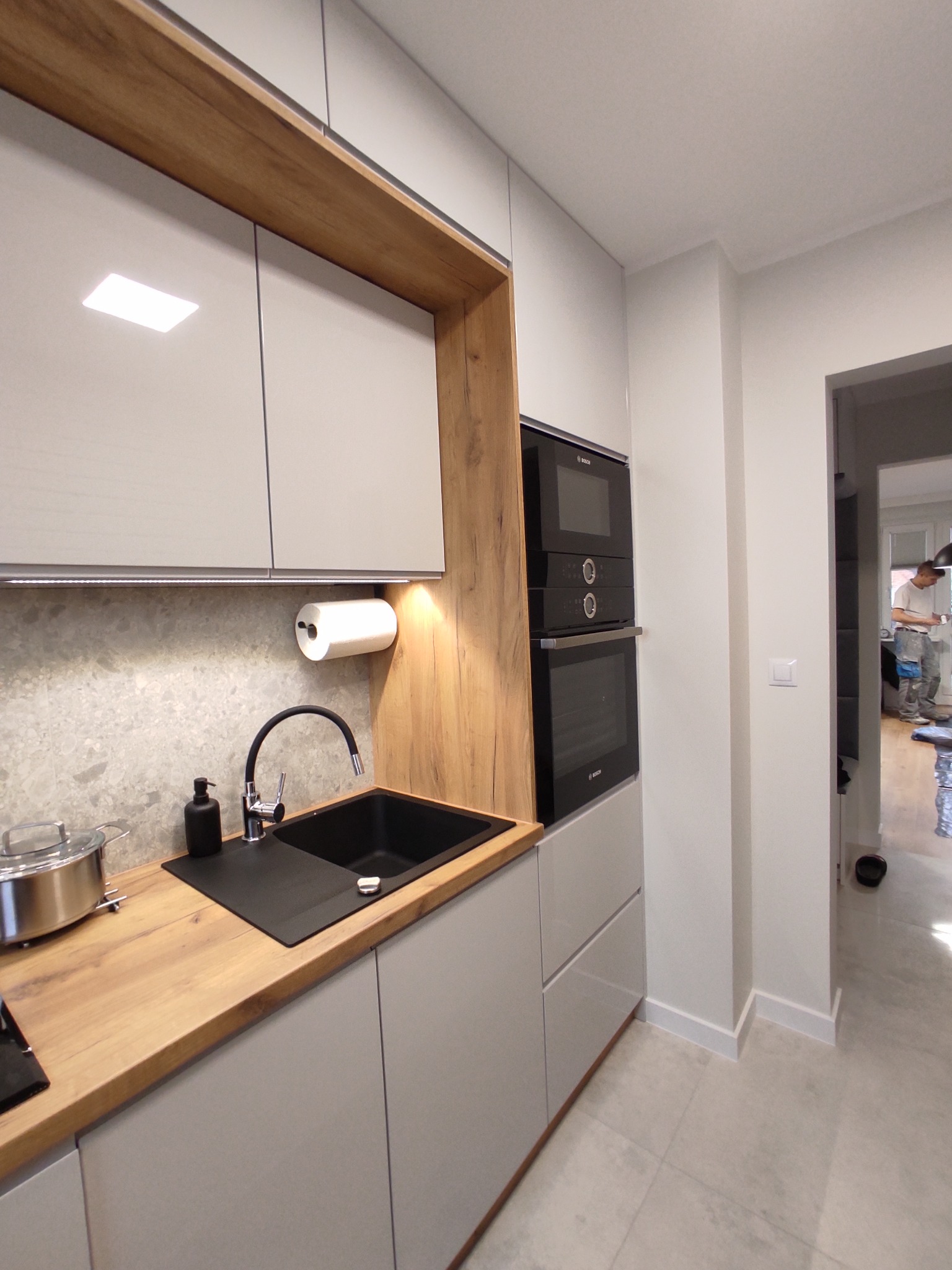 Nowoczesna kuchnia z szarymi szafkami, drewnianym blatem, czarnym zlewozmywakiem i wbudowanym piekarnikiem Bosch, widok na korytarz z pracującym malarzem.