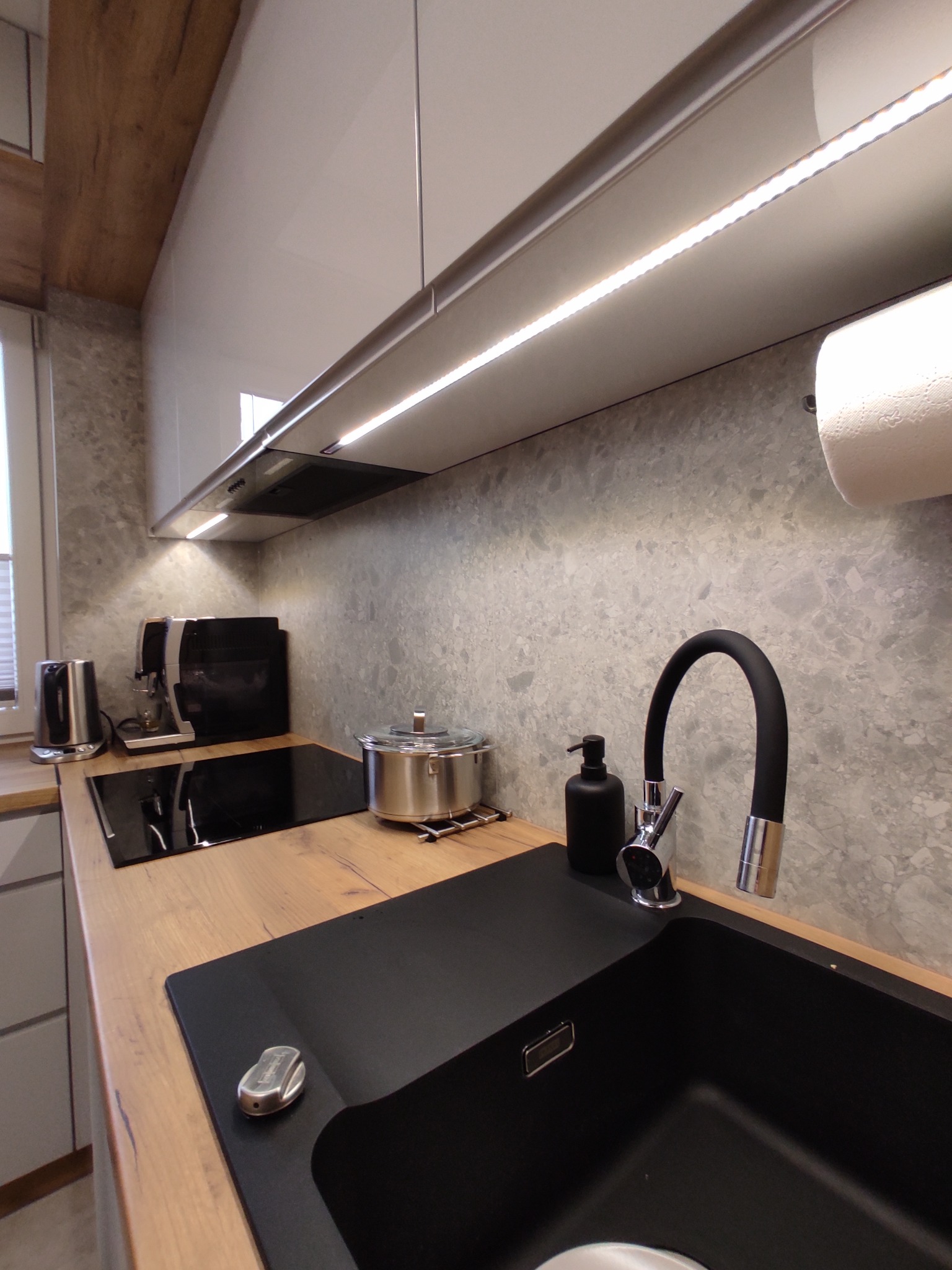 Nowoczesna kuchnia z czarnym zlewem, baterią z elastyczną wylewką, blatem imitującym drewno i szarymi ścianami z efektem kamienia, oświetlona listwą LED pod szafkami.