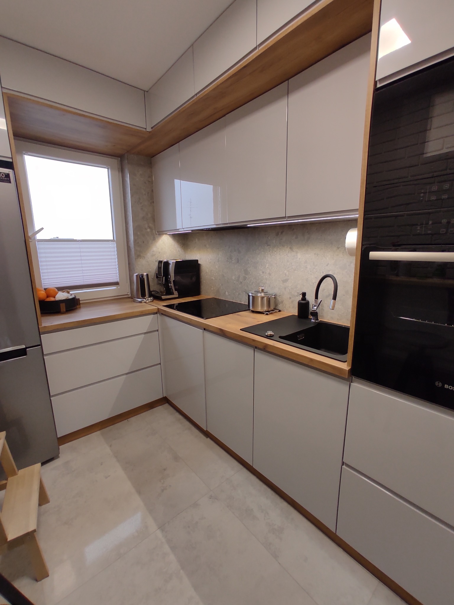 Nowoczesna kuchnia z białymi, gładkimi szafkami, drewnianym blatem i wykończeniami, czarnym zlewem i baterią, zintegrowanym piekarnikiem Bosch, ekspresami do kawy i owocami w misce przy oknie.
