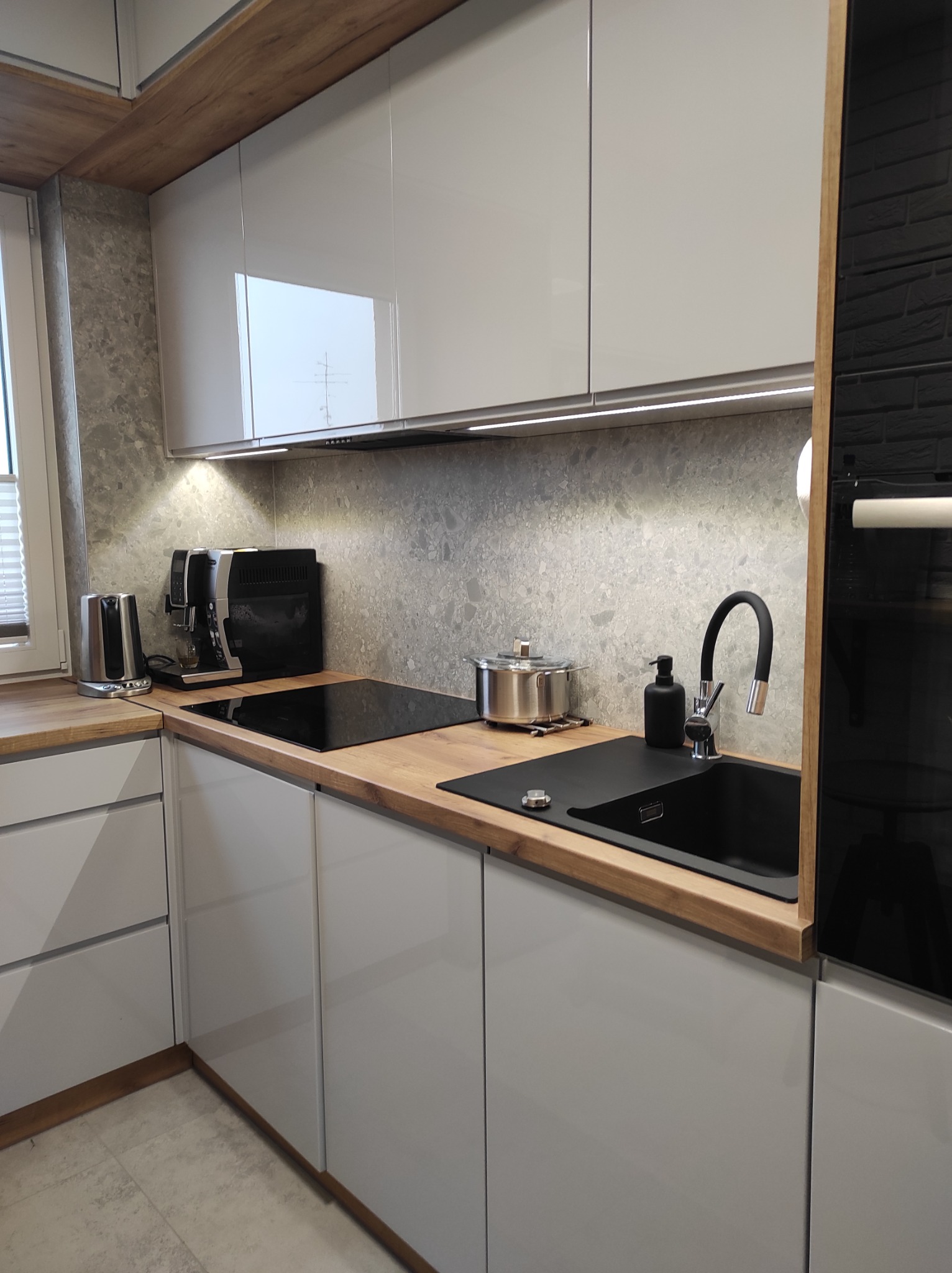 Nowoczesna kuchnia z szarymi szafkami, drewnianym blatem, czarnym zlewem i armaturą, oraz ekspres do kawy na blacie.