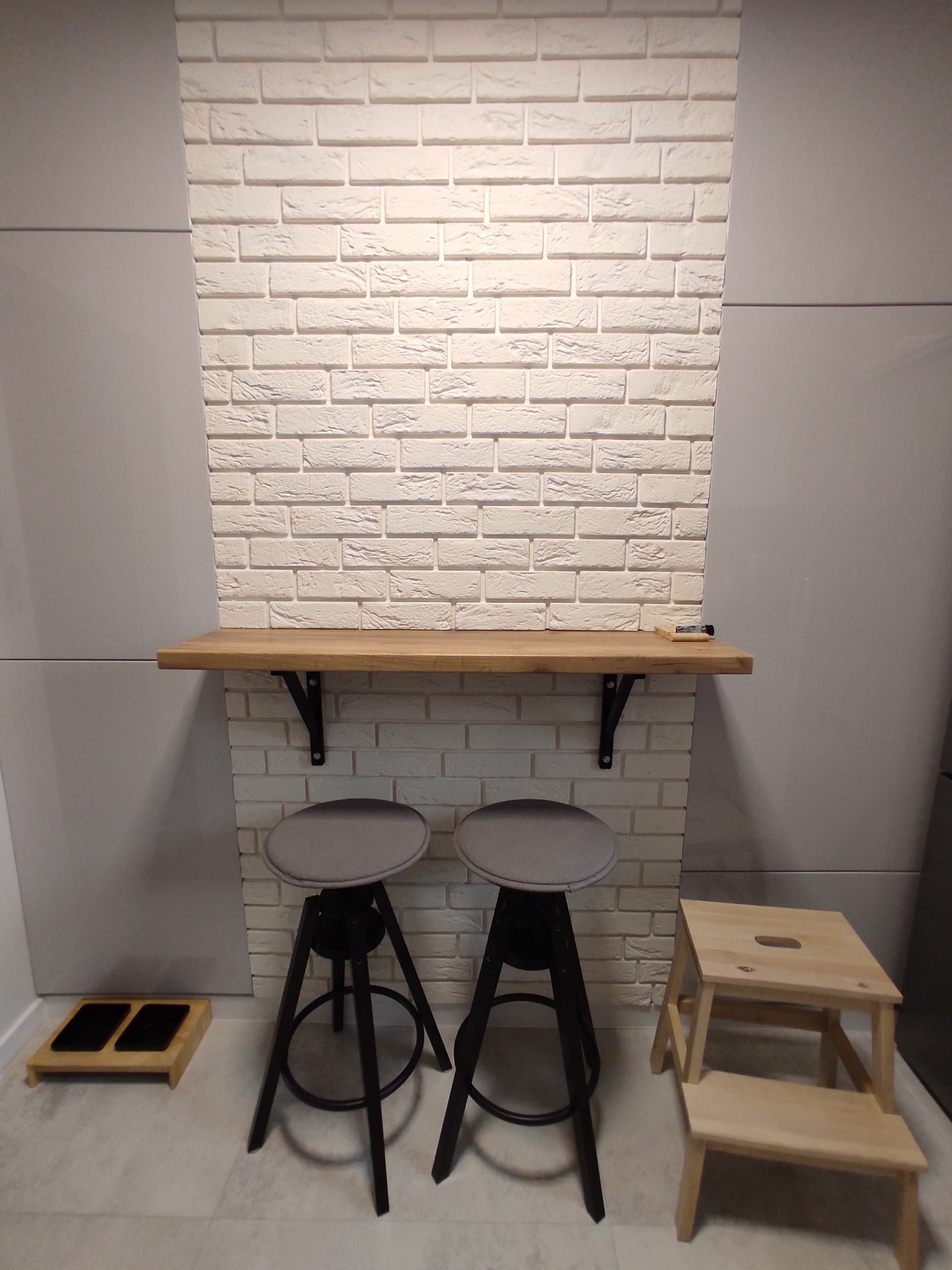Ściana z białej cegły dekoracyjnej w aneksie kuchennym, z drewnianym blatem wspartym na czarnych wspornikach, dwa hokery z szarymi siedziskami, jasne panele ścienne po bokach.