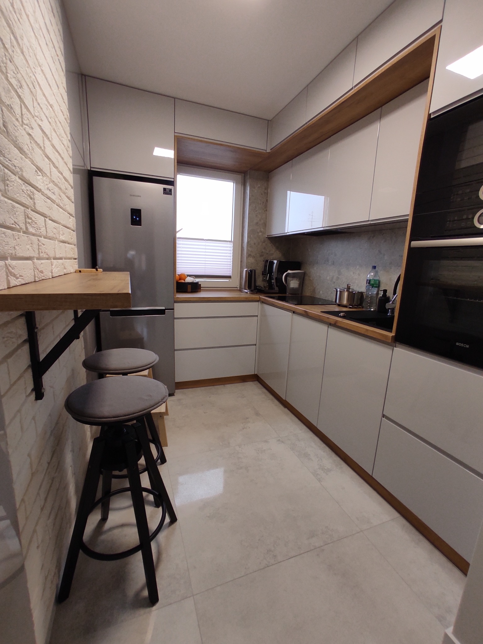 Nowoczesna kuchnia z białymi, połyskującymi szafkami, drewnianym blatem i barkiem przymocowanym do ściany z cegły, uzupełniona dwoma ciemnymi stołkami barowymi.