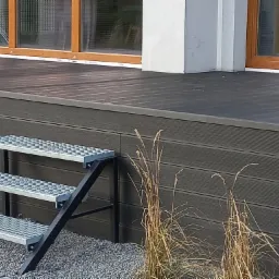 Ciemnoszary taras kompozytowy z metalowymi schodami wejściowymi i ozdobnym żwirkiem, widok z poziomu gruntu.