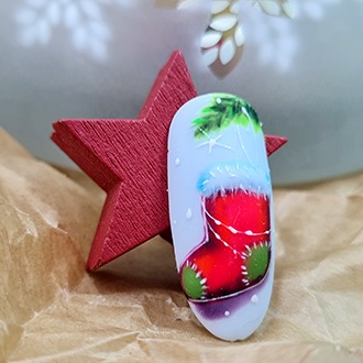 Pojedynczy paznokieć z zimową stylizacją: ręcznie malowany czerwony świąteczny but z białym futerkiem i zielonymi akcentami na jasnym tle, obok czerwona drewniana gwiazda, na tle papieru.