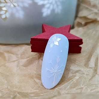 Paznokieć z zimowym zdobieniem: biały lakier, wypukły wzór śnieżynki i drobne, błyszczące elementy, na tle czerwonej gwiazdki i szarego wazonu na papierze.