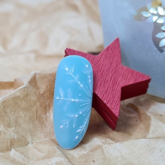 Błękitny paznokieć z ręcznie malowanym białym wzorem śnieżynki, ułożony obok czerwonej, drewnianej gwiazdki na tle zmiętego, brązowego papieru.