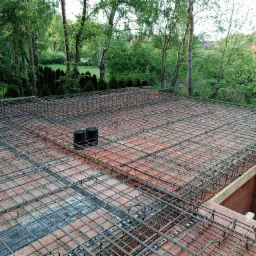 Zbrojenie siatką stalową na bloczkach fundamentowych, widoczne dwa czarne pionowe rury, w tle zieleń drzew