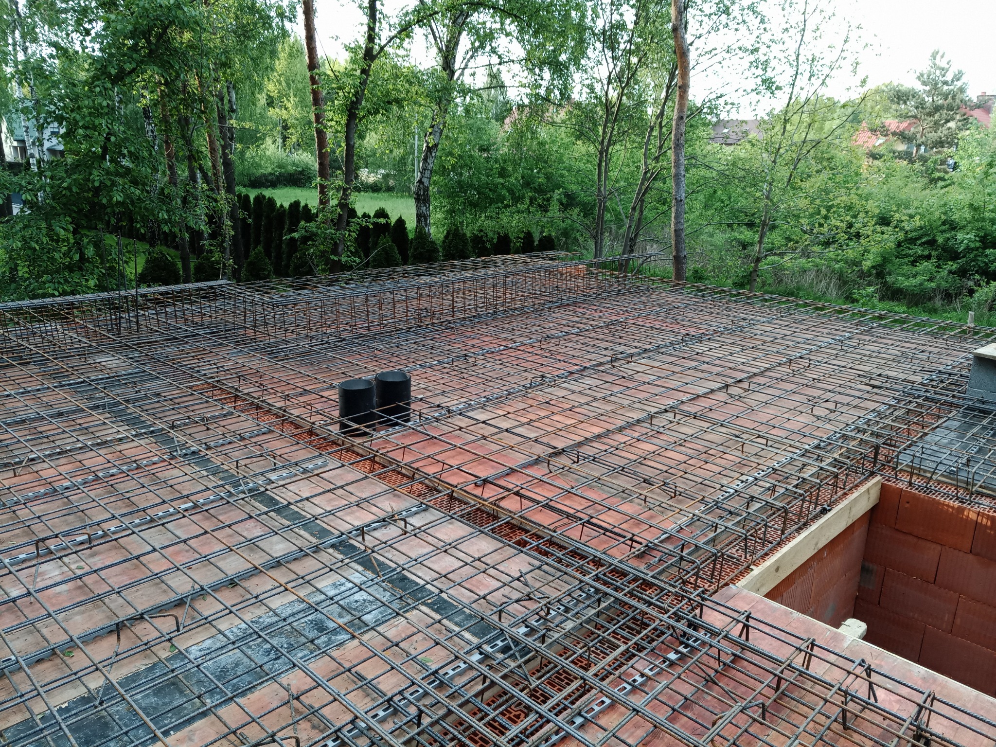 Zbrojenie siatką stalową na bloczkach fundamentowych, widoczne dwa czarne pionowe rury, w tle zieleń drzew