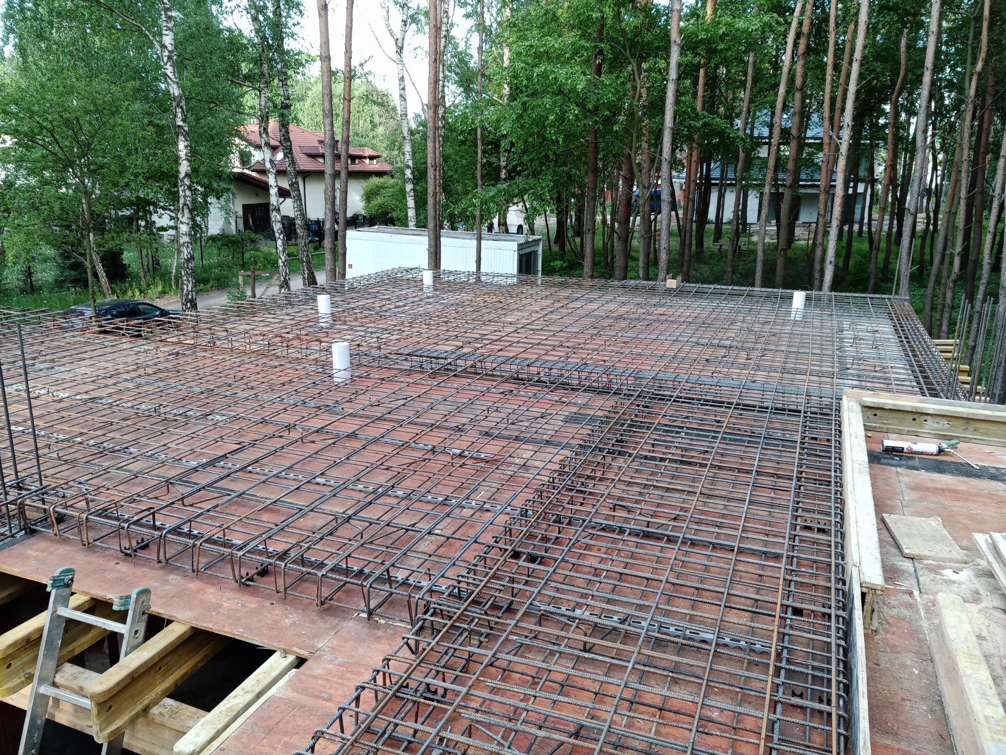 Siatka zbrojeniowa ułożona na deskowaniu, przygotowana do zalania betonem, z widocznymi rurkami pionowymi i fragmentem drabiny w tle, na tle drzew i budynku w oddali.