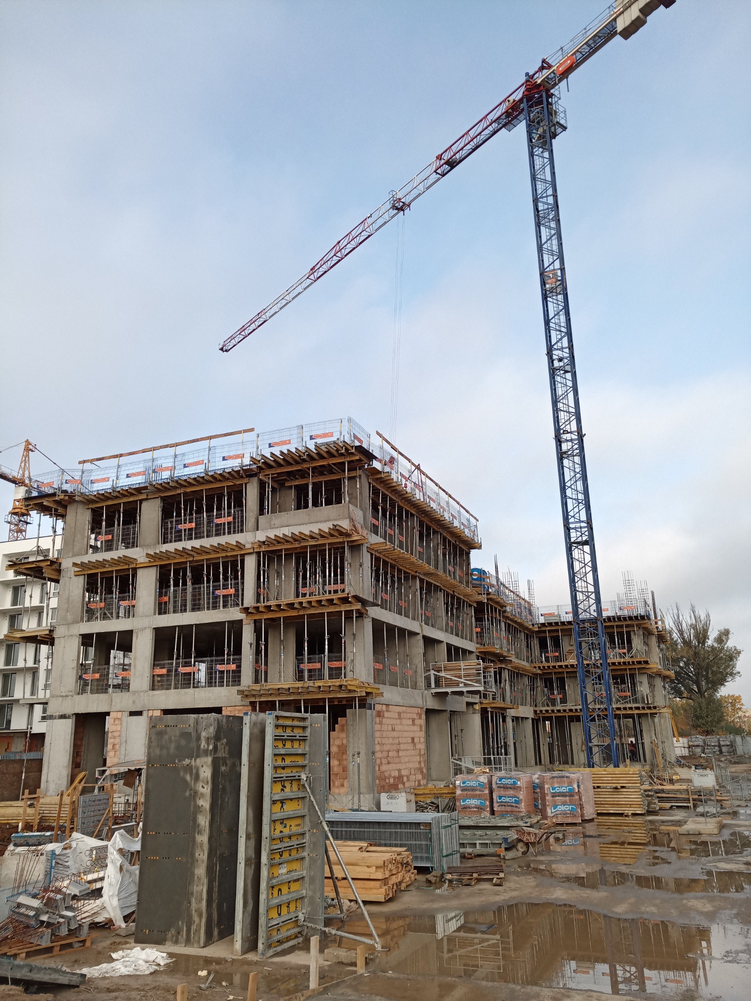 Wielopiętrowy budynek w stanie surowym, z widocznymi szalunkami i zbrojeniami, na pierwszym planie stojące elementy szalunkowe, w tle wysoki żuraw budowlany, po deszczu.