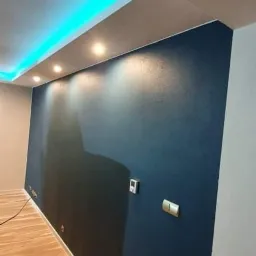 Wykończona ściana w odcieniach granatu z włączonymi kontaktami, oświetlenie sufitowe LED i halogenowe, drewniana podłoga oraz fragment schodów z drewnianą balustradą.