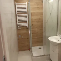 Zakończona realizacja: nowoczesna łazienka z kabiną prysznicową, grzejnikiem drabinkowym i umywalką z szafką, wykończona płytkami imitującymi drewno, oświetlona światłem dziennym.