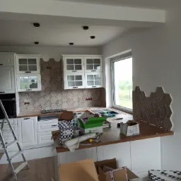 Montaż heksagonalnych płytek na ścianie w kuchni, widoczne puszki elektryczne, białe szafki z przeszklonymi frontami, drabina i kartony z materiałami budowlanymi na blacie.