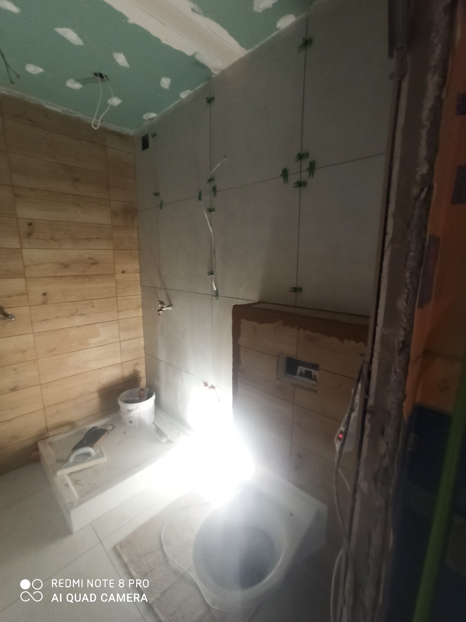 Łazienka w trakcie układania płytek: jasne płytki na ścianie z krzyżykami dystansowymi, drewnopodobne płytki na drugiej ścianie, widoczne narzędzia i sanitariat, sufit z płyt kartonowo-gipsowych.