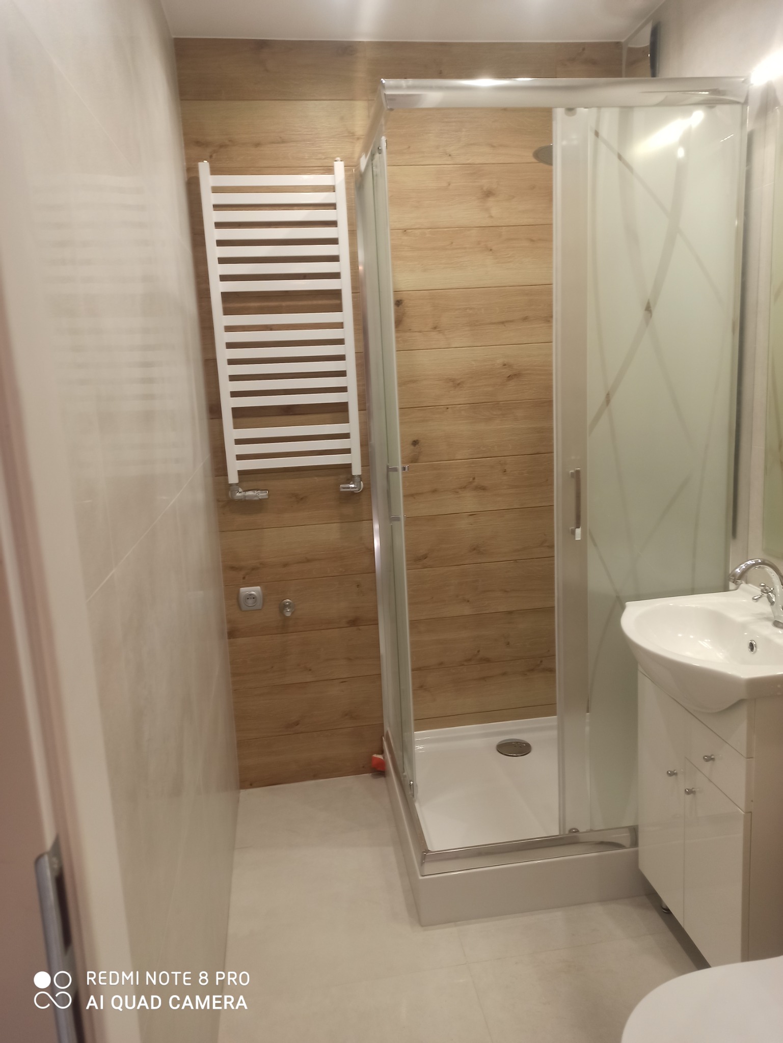 Zakończona realizacja: nowoczesna łazienka z kabiną prysznicową, grzejnikiem drabinkowym i umywalką z szafką, wykończona płytkami imitującymi drewno, oświetlona światłem dziennym.