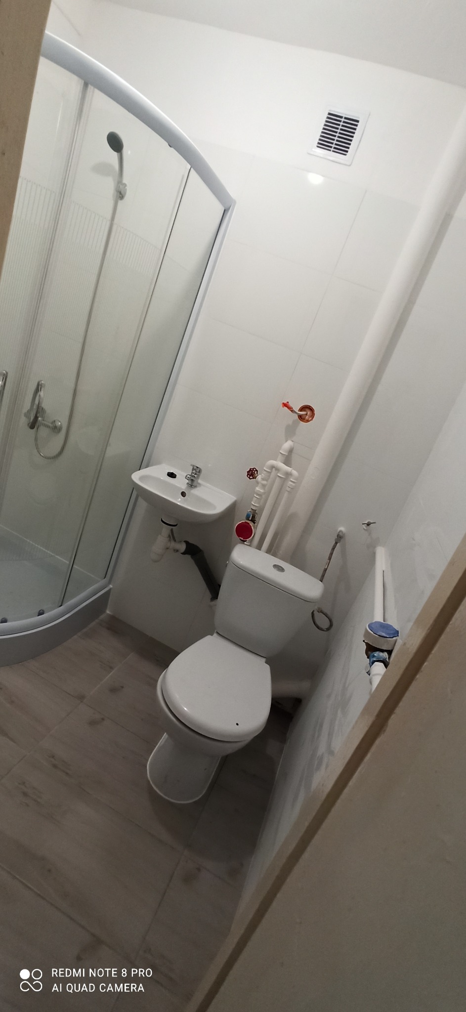 Ukończona łazienka z białą kabiną prysznicową, umywalką i toaletą, widoczne rury i wentylacja, płytki w odcieniach szarości i bieli.