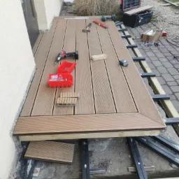 Montaż tarasu kompozytowego na regulowanych wspornikach, widoczne narzędzia, skrzynka narzędziowa, elementy mocujące i fragment kostki brukowej.