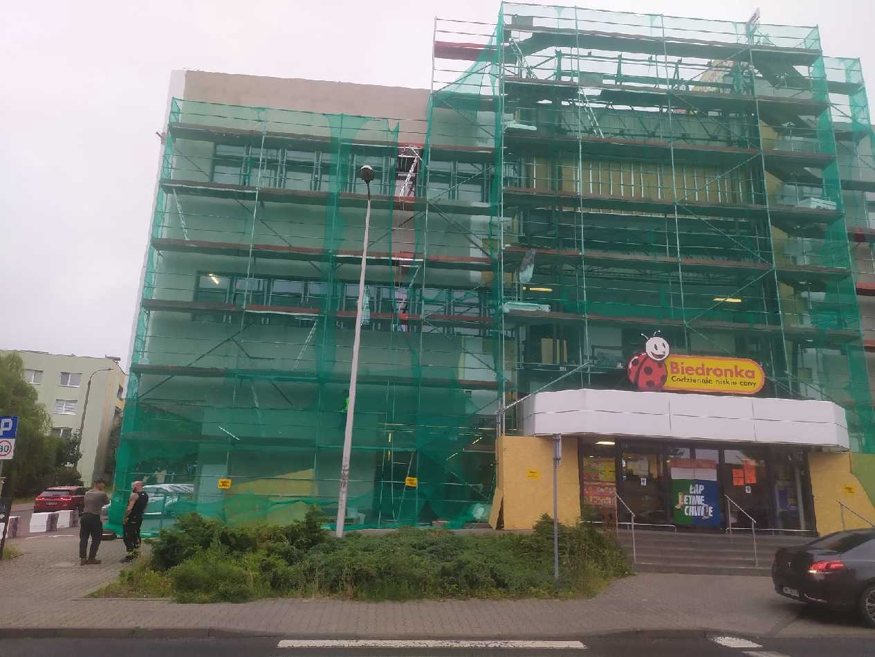 Budynek sklepu Biedronka w trakcie remontu elewacji, osłonięty rusztowaniami i siatką ochronną. Widoczni pracownicy oraz zaparkowany samochód.