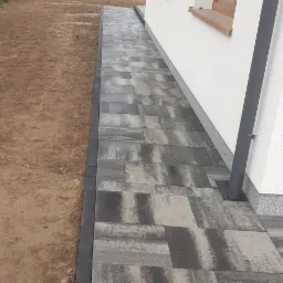 Świeżo ułożony chodnik z szarych, prostokątnych płyt betonowych obramowany ciemnymi kostkami, biegnący wzdłuż białej ściany budynku z drewnianymi oknami.