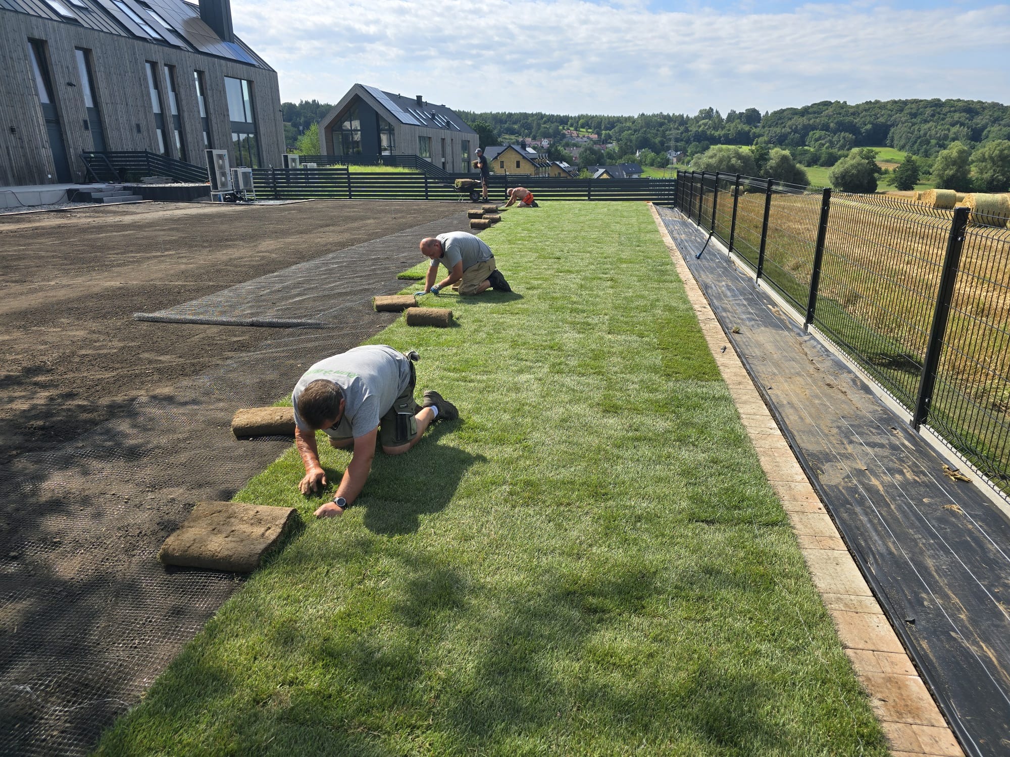 Ekipa montuje świeżą darń na przygotowanym terenie, nowoczesny dom w tle, widoczne rolki trawy i siatka wzmacniająca podłoże.
