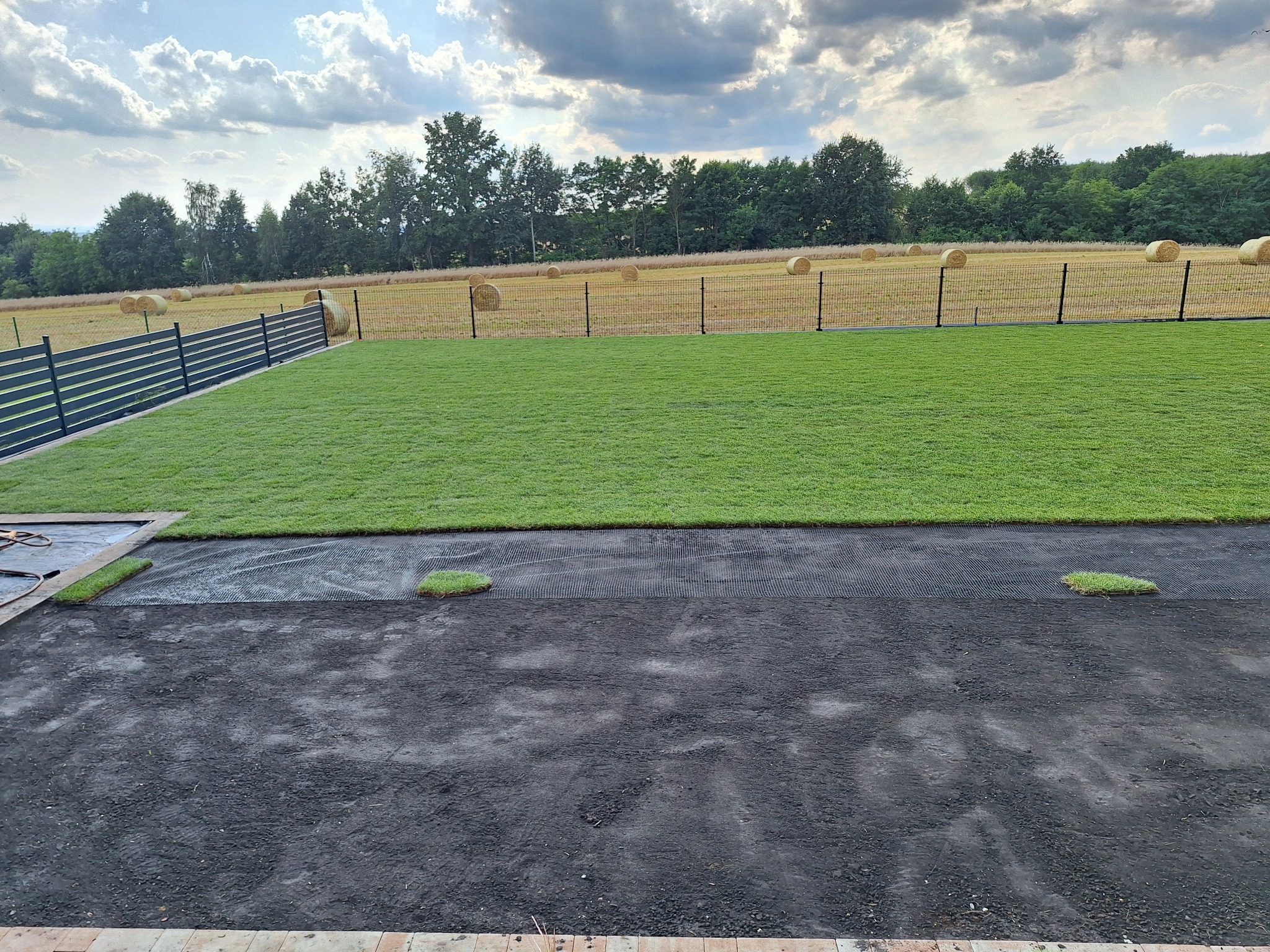 Fragment nowo założonego trawnika z rolki, widoczne łączenia i przygotowany teren, w tle pole ze słomianymi balotami i nowoczesne ogrodzenie.