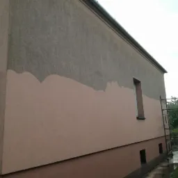 Elewacja budynku w trakcie malowania; górna część szara, dolna część w odcieniach różu, widoczne rusztowanie i rynna z numerem 15.