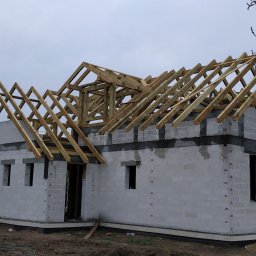 A-Z Adam Zdanowski - Surowy stan domu w trakcie budowy z pustaków, z widoczną drewnianą konstrukcją dachu na tle pochmurnego nieba.