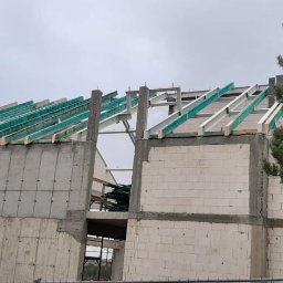 A-Z Adam Zdanowski - Wznoszenie konstrukcji domu: surowe ściany z betonu komórkowego, stalowe słupy i drewniane krokwie z zieloną folią zabezpieczającą, widok z zewnątrz na tle pochmurnego nieba.