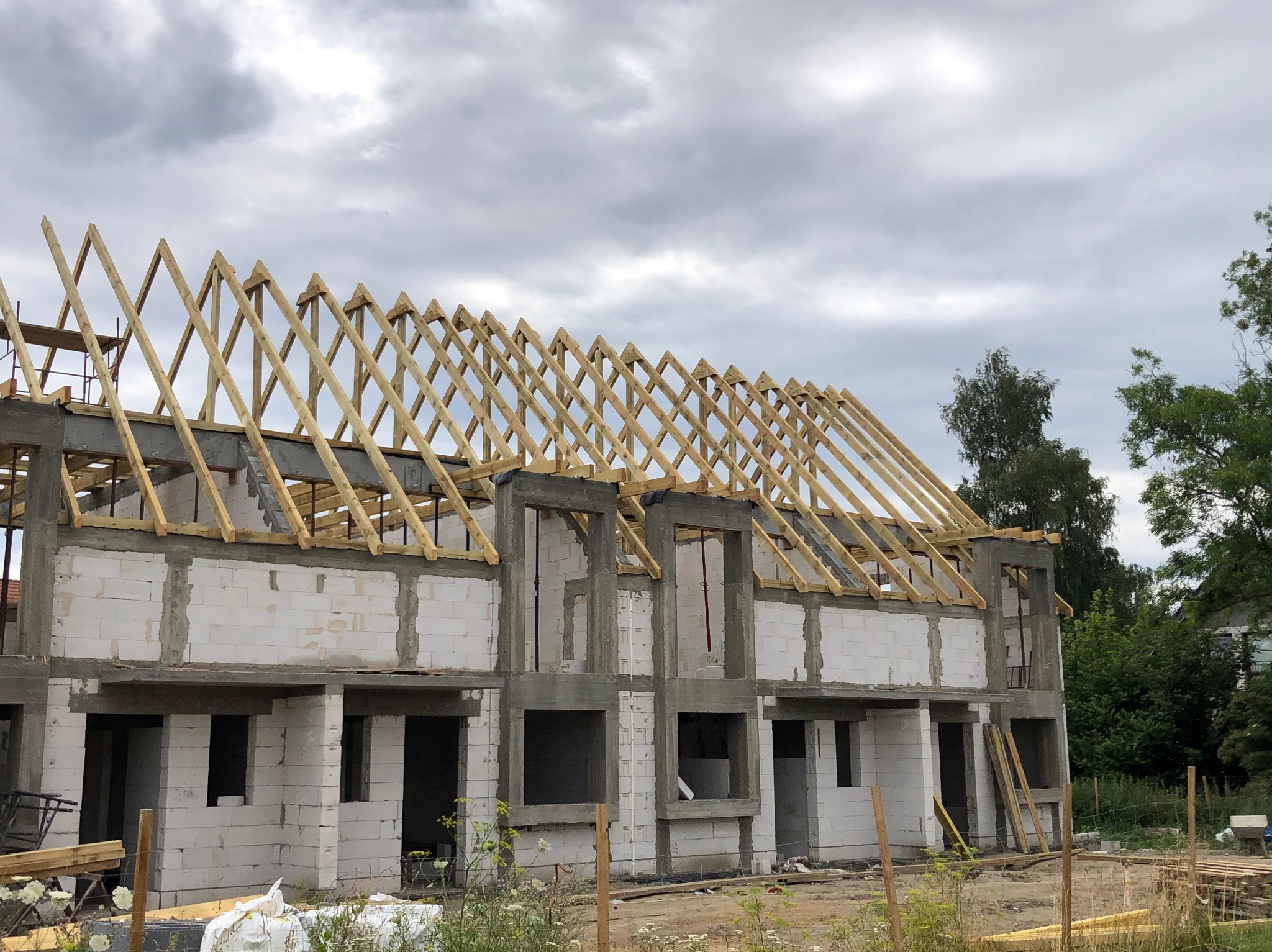 Surowy stan budowy domu: ściany z białych bloczków, betonowe nadproża i drewniana konstrukcja dachu pod pochmurnym niebem.