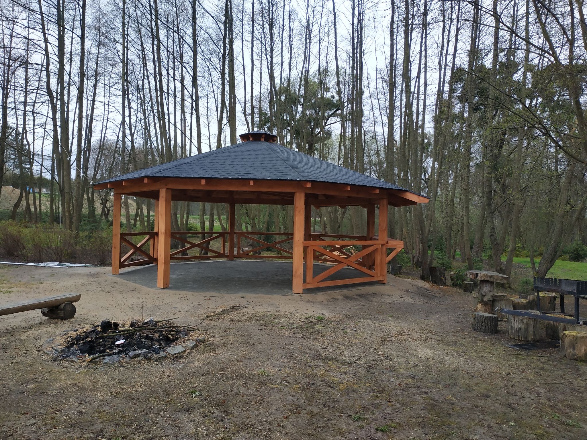 Drewniana konstrukcja z czarnym, wielospadowym dachem, z ozdobnymi krzyżowymi belkami, otoczona drzewami, obok palenisko i siedziska z pniaków.