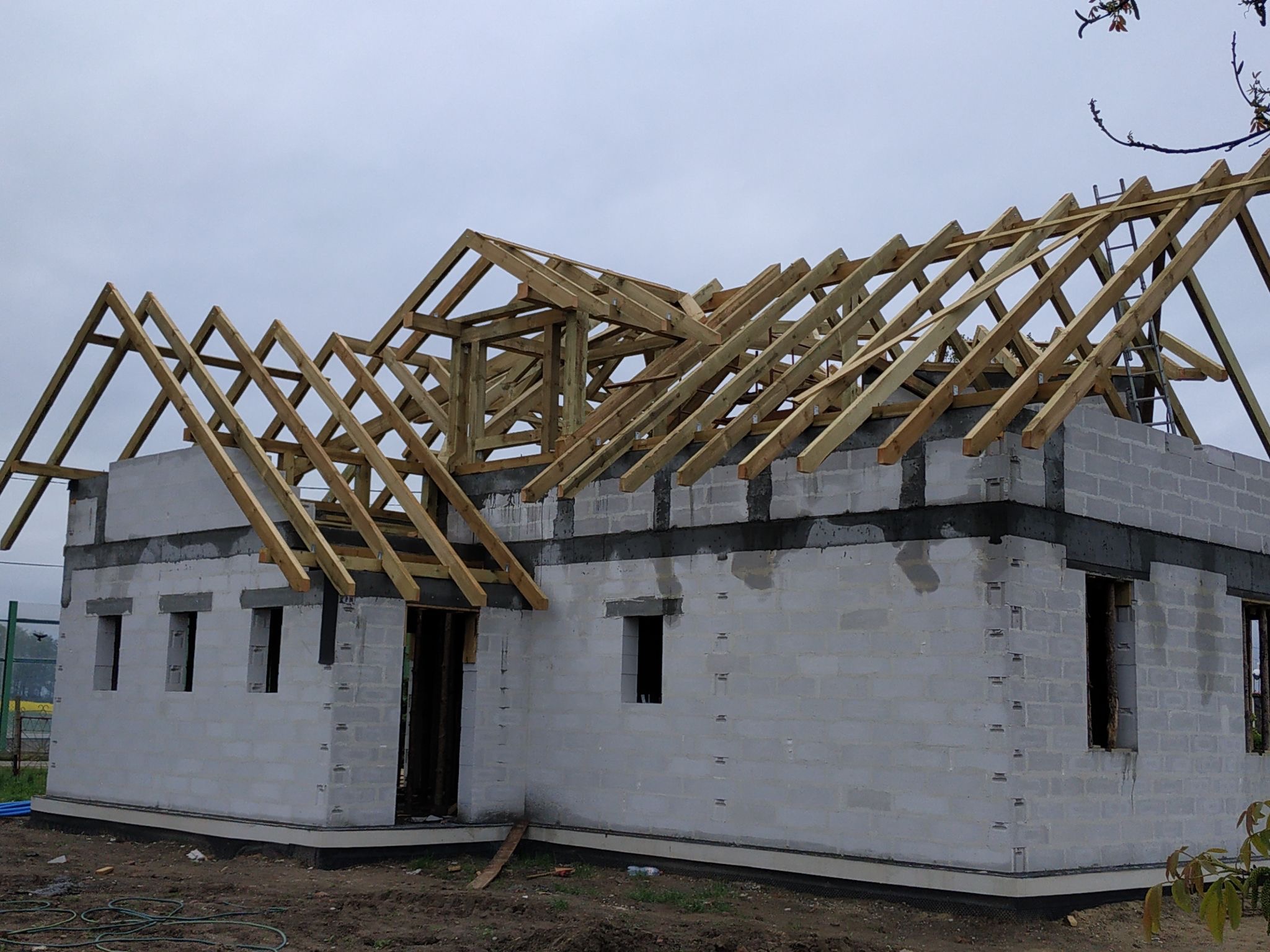 Surowy stan domu w trakcie budowy z pustaków, z widoczną drewnianą konstrukcją dachu na tle pochmurnego nieba.