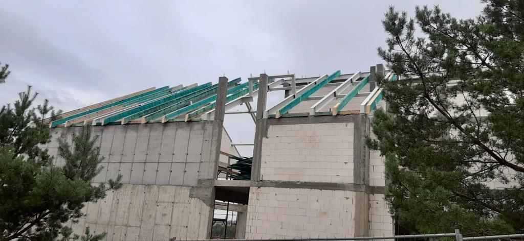 Wznoszenie konstrukcji domu: surowe ściany z betonu komórkowego, stalowe słupy i drewniane krokwie z zieloną folią zabezpieczającą, widok z zewnątrz na tle pochmurnego nieba.