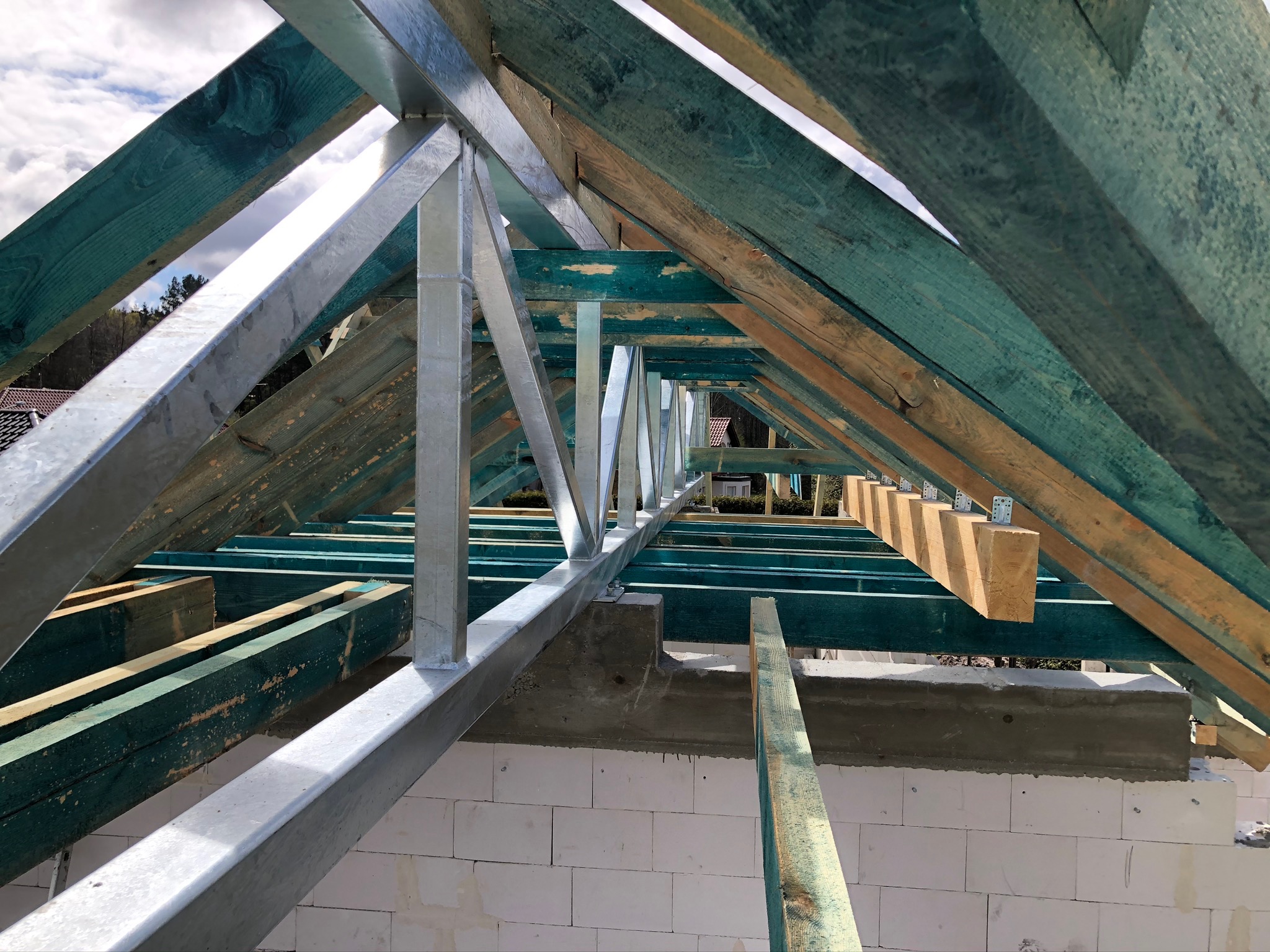 Konstrukcja dachu z elementami stalowymi i drewnianymi belkami pomalowanymi na zielono, widok od wewnątrz, białe bloczki fundamentowe w tle.