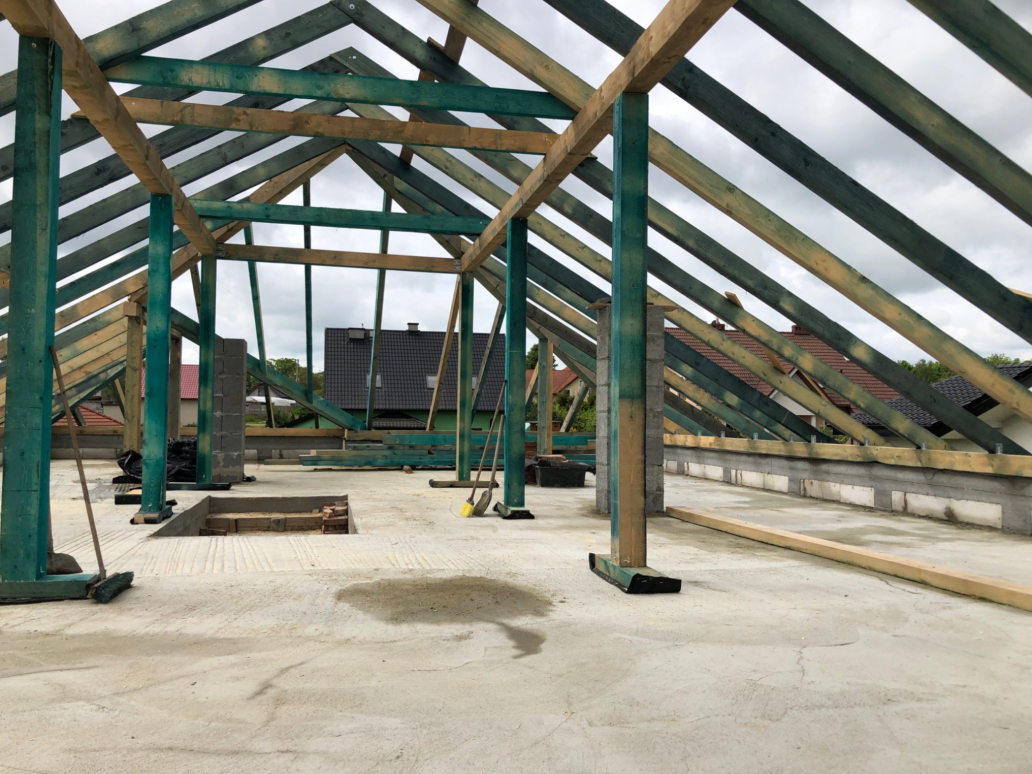 Wnętrze w trakcie budowy domu, widoczny szkielet dachu z impregnowanego drewna, betonowa podłoga z otworami, w tle domy jednorodzinne.