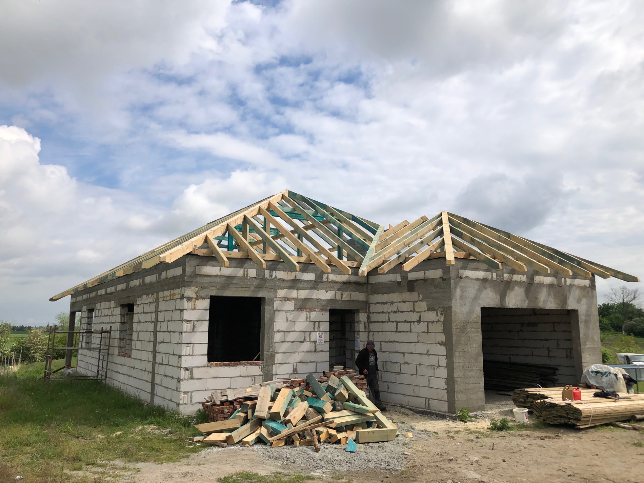 Budowa domu z pustaków: surowy stan z drewnianą konstrukcją dachu, stos pociętych desek i pracownik w tle.
