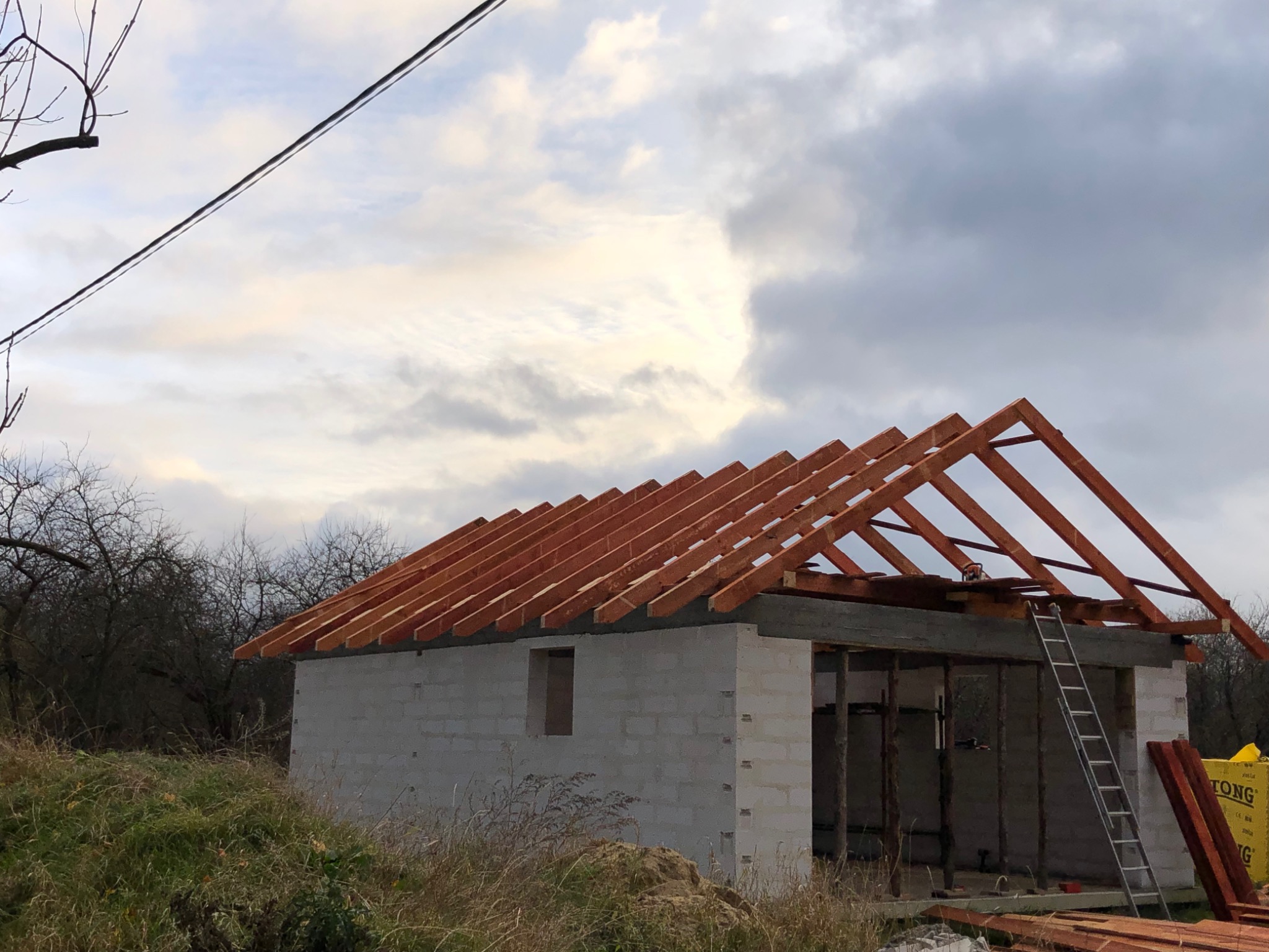 Budowa parterowego budynku z bloczków, z widoczną konstrukcją dachu z drewnianych belek, drabina oparta o ścianę, w tle pochmurne niebo.