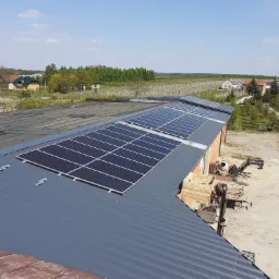 Panele słoneczne na szarym dachu budynku gospodarczego, widok z góry, w tle zabudowania wiejskie i sad.