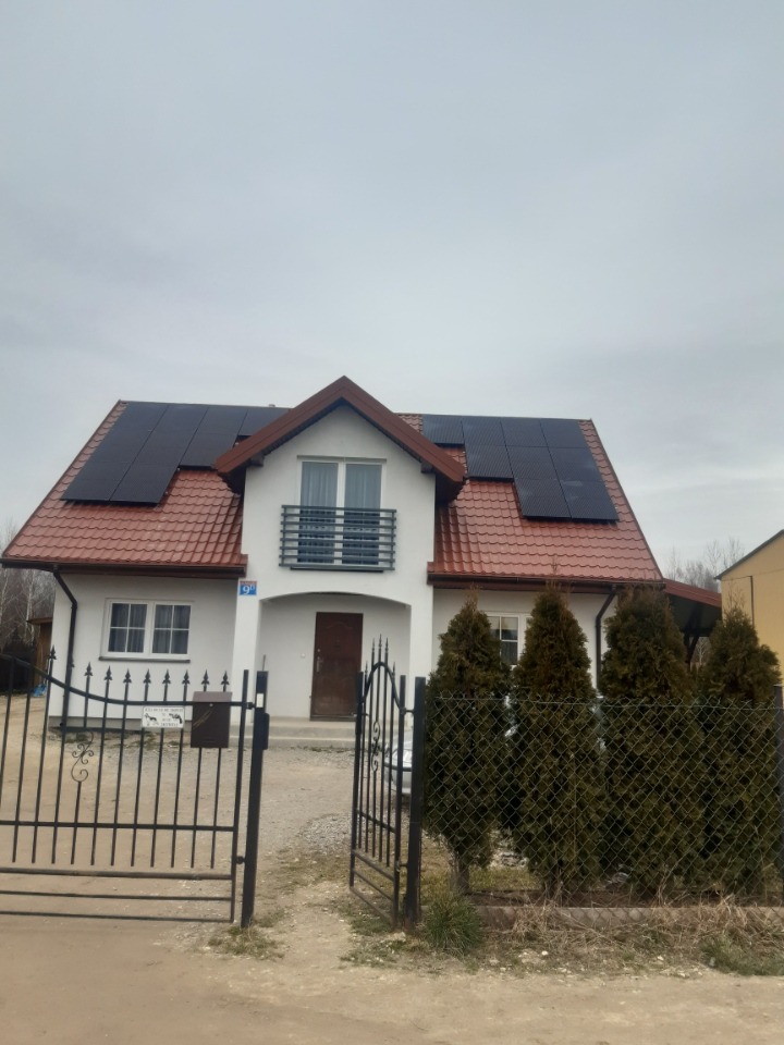 Dom jednorodzinny z zainstalowanymi panelami słonecznymi na dachu pokrytym czerwoną dachówką, widoczne przez otwartą, metalową bramę.