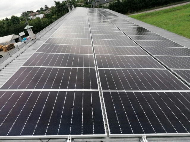 Rząd paneli słonecznych na płaskim, szarym dachu z widocznym kominem wentylacyjnym i zielenią w tle. Panele ustawione w rzędach, z odbiciami nieba na powierzchni.