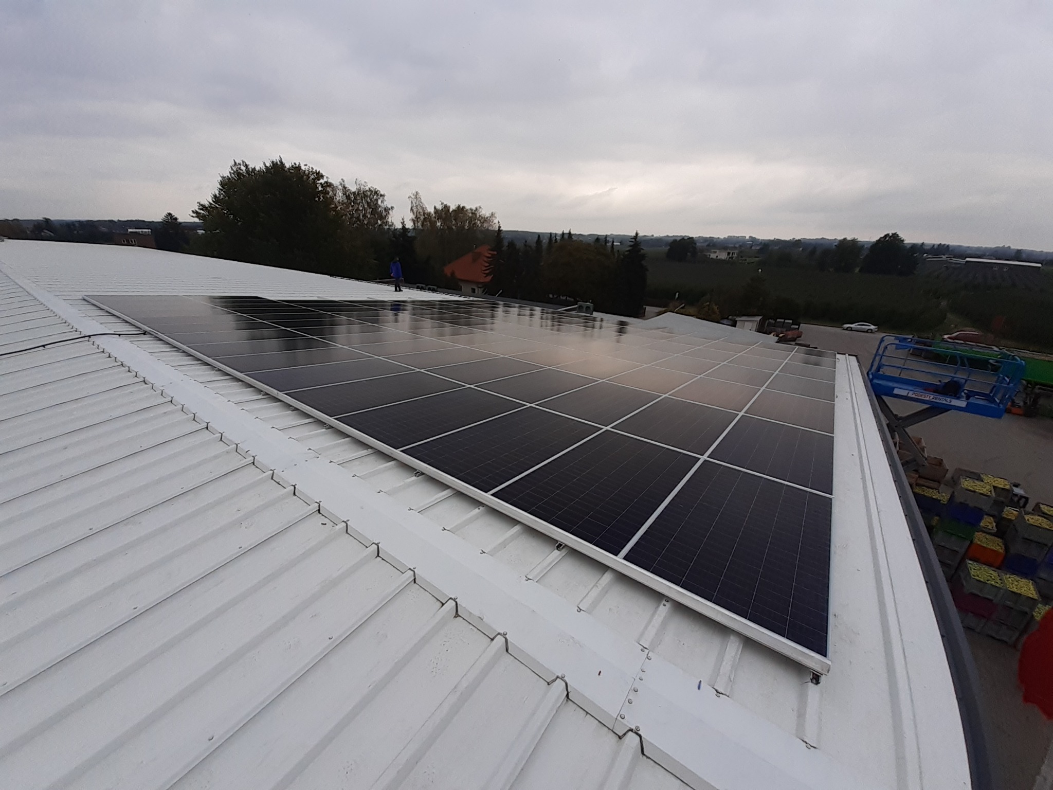 Rząd paneli słonecznych na białym, metalowym dachu budynku przemysłowego, z widoczną osobą w niebieskiej kurtce i okolicznym krajobrazem w tle.