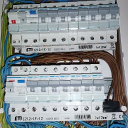 Rozdzielnica elektryczna z zamontowanymi bezpiecznikami Hager i ETI, widoczne kable w kolorach niebieskim, brązowym i żółto-zielonym, podłączone do listew zaciskowych.