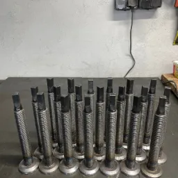 Partia metalowych śrub z gwintem trapezowym i kwadratowym zakończeniem, ułożonych rzędami na metalowym stole warsztatowym; widoczne ślady spawania i obróbki.