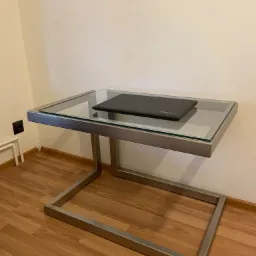 Stolik kawowy ze szklanym blatem i metalową ramą w kształcie litery U, na blacie leży laptop Lenovo.
