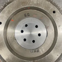 Metalowa zębatka koła zamachowego z sześcioma otworami w centralnej części, widoczne ślady użytkowania i drobne rysy na powierzchni.