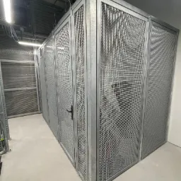 Szereg metalowych, siatkowych boksów zamykanych na czarne klamki, umieszczonych w hali przemysłowej z widoczną instalacją pod sufitem i roletą w tle.